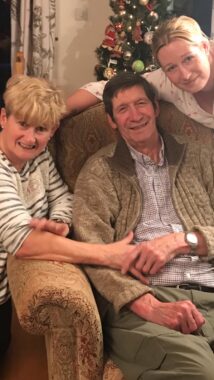 A family photo in front of a Christmas tree, with the father sitting in a large chair with his wife at his right (our left) and his daughter to his left (our right). All are smiling. 