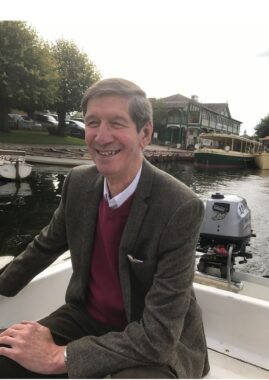 An older English gentleman smiles while navigating a small fishing boat on a river. He's dressed in a pink dress shirt covered by a red sweater and a tweed jacket. He looks happy despite the typical overcast skies of England.