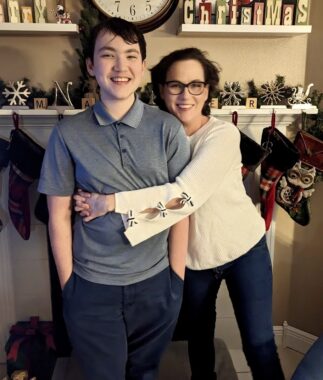 A woman embraces a teenage boy in front of a mantel with christmas decorations.