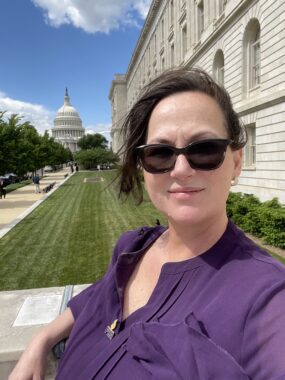 A woman in a purple blouse takes a selfie on a sunny day in Washington, D.C., with the U.S. Capitol visible in the background. 