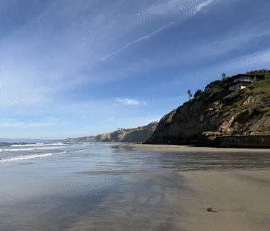 A postcard-like beach photo on the rugged California coast. Low waves roll in during low tide, and the skies are mostly clear and light blue. 