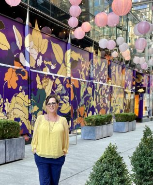 A woman wearing a yellow top and blue slacks smiles for a photo in an outdoor walkway. The wall behind her depicts a colorful mural, and there are pink and purple lanterns strung up over the walkway.