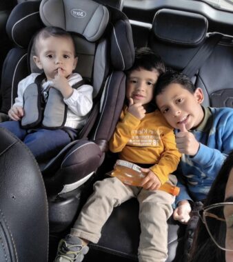 Three young children are scrunched together in the back seat of a car. The youngest, to the left, is in a car seat. The other two are flashing a thumbs up. 