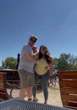 A man and woman pose for a photo with their newborn baby. They're standing outside on some sort of patio and it looks to be a warm, sunny day.
