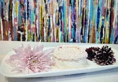 A small, white, frosted cake sits between two decorative flowers on a long serving platter. A colorful, abstract painting of trees hangs in the background.