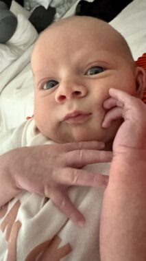 A 4-week-old baby holds his hands up near his face and looks curiously at the camera.