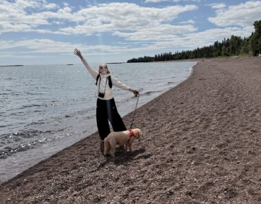 A woman waves as she stands with a dog by the shore of a lake.
